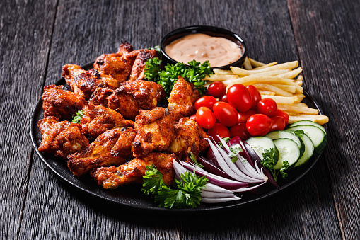 crispy grilled chicken wings with french fries, tomatoes, cucumber, red onion and thousand islands dip on black plate on dark wood table