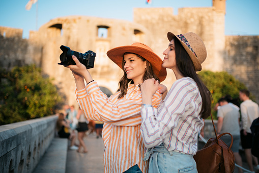 Two happy female tourists walking around and exploring the port city of Dubrovnik and taking photographs of an old architecture with a professional photographic camera