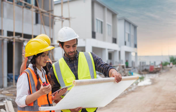 hombres caucásicos y mujeres asiáticas constructoras, arquitectas e ingenieras con un borrador de plan de construcción y computadora portátil hablando sobre el sitio de construcción - building engineering fotografías e imágenes de stock