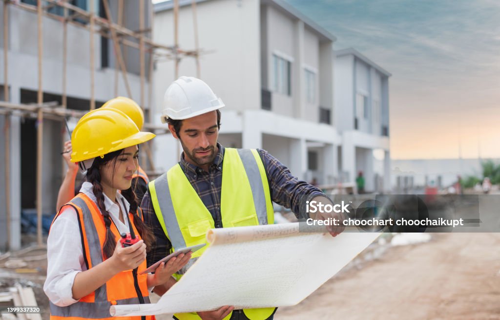 Caucasian males and Asian female builders, architector and engineers with draft plan of building and laptop computer talking on constructing site Engineer Stock Photo