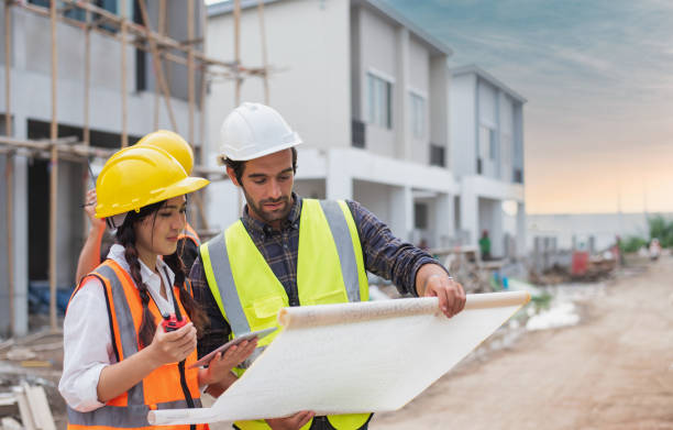 Caucasian males and Asian female builders, architector and engineers with draft plan of building and laptop computer talking on constructing site stock photo