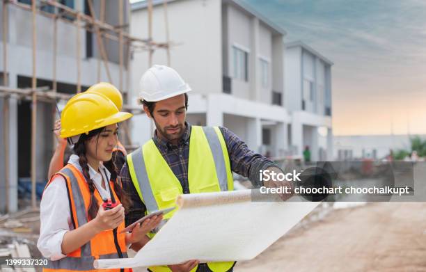 Caucasian Males And Asian Female Builders Architector And Engineers With Draft Plan Of Building And Laptop Computer Talking On Constructing Site Stock Photo - Download Image Now