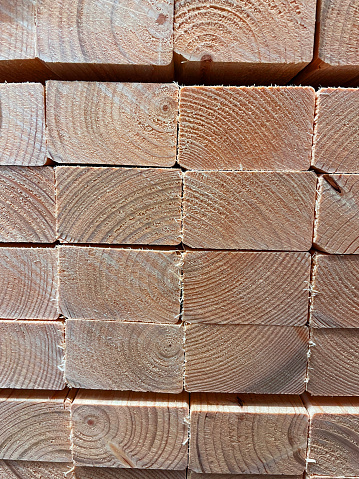 Stock photo showing close-up view of pile of rectangular sawn wooden plank lengths, which are stacked up together to form a regular background pattern. The neatly sawn ends are displaying the woodgrain and concentric age rings with their different colours and shapes.