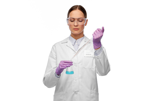 science, chemistry and people concept - young female scientist in goggles with pipette chemical in flask making test or research