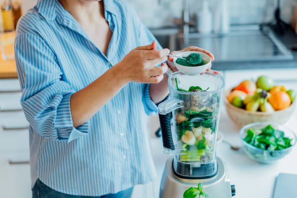 女性は台所でグリーンスムージーを作る際にスピルリナグリーンパウダーを追加します。スーパーフードサプリメント。健康的なデトックスビーガンダイエット。健康的なダイエット、減量� - スーパーフード ストックフォトと画像
