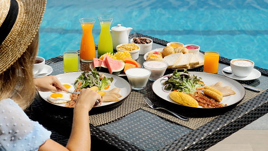 Buffet service. Tasty breakfast served on table. Travel woman in hat eating breakfast is served with eggs, sausage, coffee, fresh orange juice, croissants, exotic fruits. Balanced diet on vacation