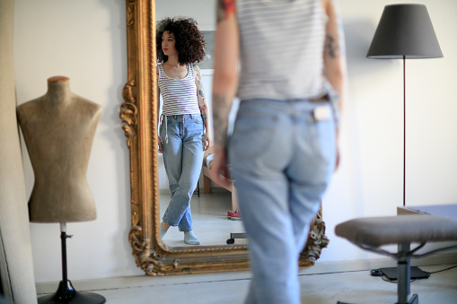 Young tattooed woman with curly hair standing in front of the old-fashioned mirror, standing cross-legged and looking away