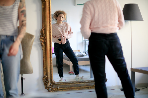Young woman with friend trying on her clothes in front of an old-fashioned mirror at home, tying sweater in front and trying new style