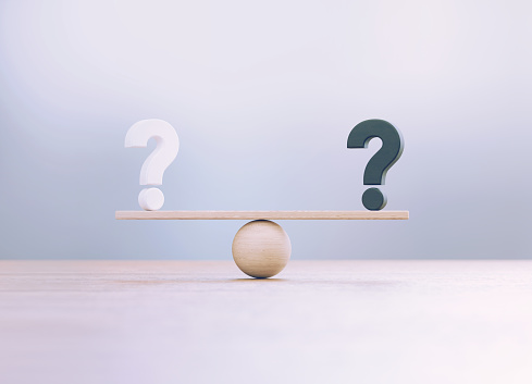 White And Black Question Marks Sitting Over Wooden Seesaw Scale Before Defocused Background
