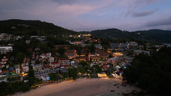 View of the beach and the island from a height. Sunset. People walk on the beach, relax. There are clouds in the sky. Green hills on the island of Phuket. There are hotels, lanterns are lit. Journey