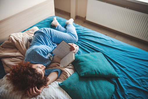 Caucasian curly-haired woman laying comfortably on bed reading a book. Curly woman reading book for fashion lifestyle design.