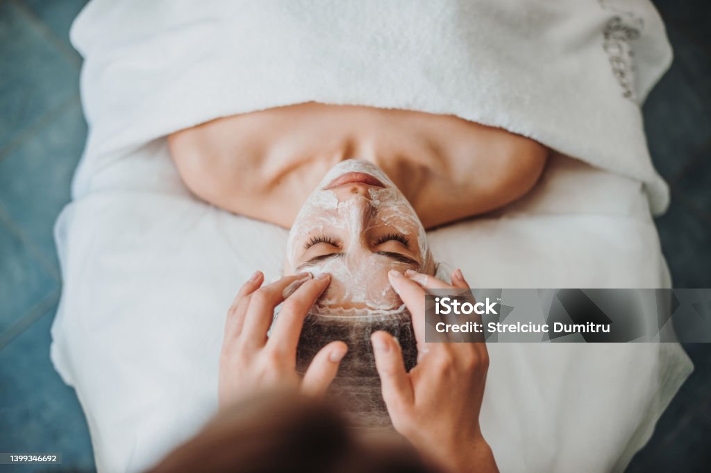 Woman getting facial care by beautician's hands at spa salon. Beautiful girl. Beauty face. Natural beauty. Cosmetology beauty procedure. Facial skin treatment. Rejuvenation treatment. Facial Mask - Beauty Product Stock Photo