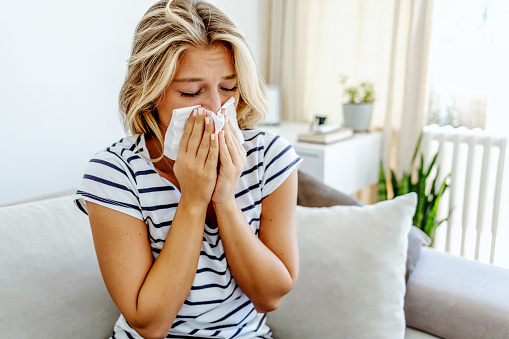 Shot of an attractive young woman feeling ill and blowing her nose with a tissue at home. Photo of sneezing woman in paper tissue. Picture showing woman sneezing on tissue on couch in the living-room.