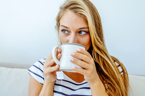Thoughtful woman sitting on couch holding a cup of tea in living room. Portrait of a young woman drinking a cup of tea while relaxing on sofa at home. Portrait of girl with hot drink in hands, looking away.