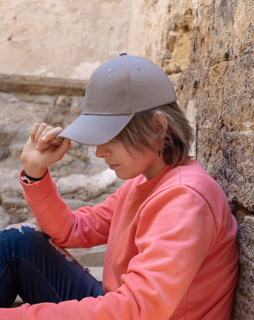 Teen girl in grey baseball cap and pink long sleeve shirt, in profile. Cap mockup. Serious Teenager girl seating near a stone wall outdoor and touching the visor. Loneliness concept
