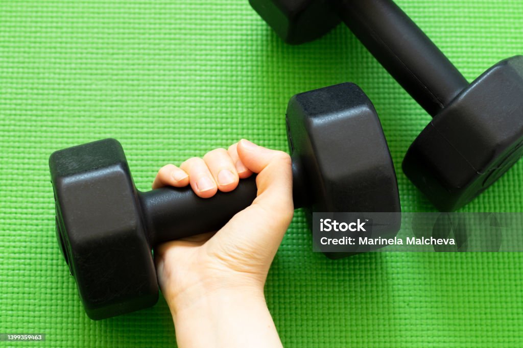Female's hand holding a dumbbell on green workout mat background Female's hand holding a dumbbell on green workout mat background. Top view, flat lay. Strong woman, lifting weights, exercise at home concept. A close-up. Barbell Stock Photo