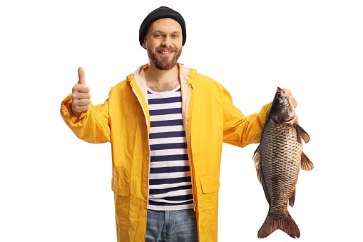 Young fisherman in a yellow raincoat holding a carp fish and gesturing thumbs up isolated on white background