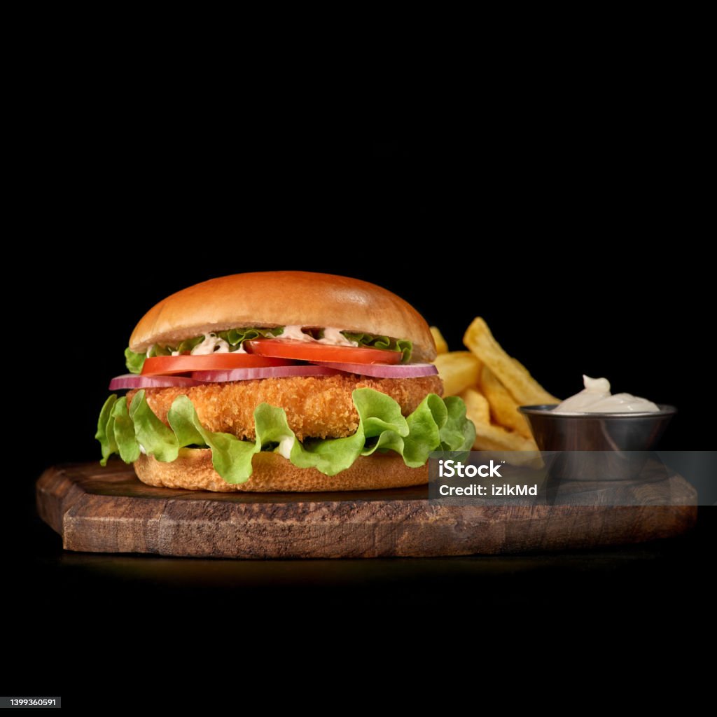 Delicious fastfood fresh tasty burger. crispy, breaded chicken burger with and vegetable on wooden cutting board Delicious fastfood fresh tasty burger. crispy, breaded chicken burger with and vegetable and french fries on wooden tray, isolated on dark background American Culture Stock Photo