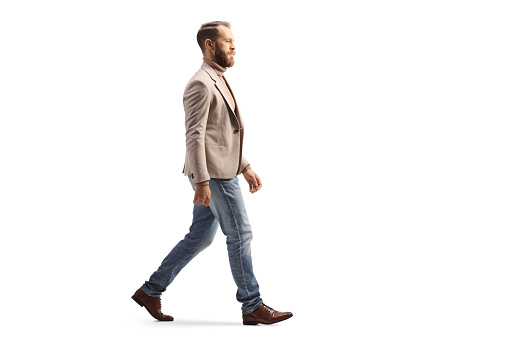 Full length profile shot of a man in a beige suit and jeans walking isolated on white background