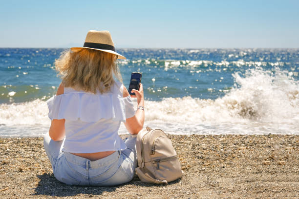 istock Woman in straw hat sitting at the sea using phone 1399362744