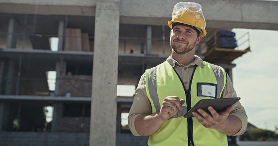 A building contractor using a digital tablet while working on a construction site. A young man browsing online with a smart device while working on an architecture project