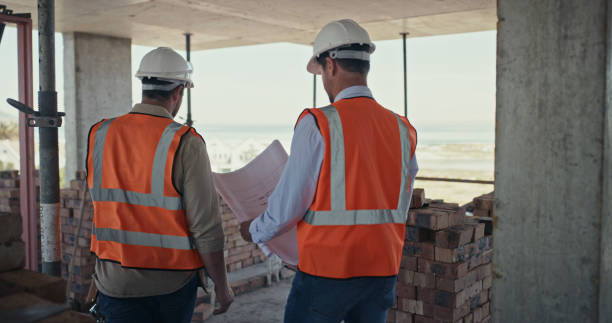 istock Builders reading a blueprint and talking at a construction site. Rear view of two men working as a team on an architecture project. Engineers and colleagues discussing plans 1399364884