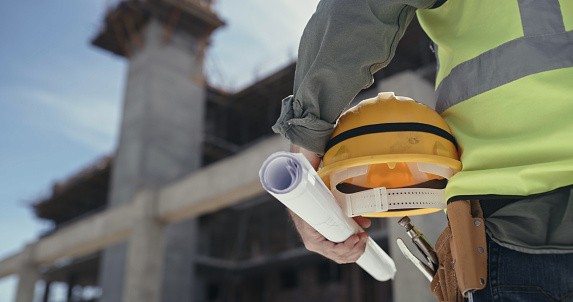 Closeup of a building contractor standing at a construction site. An unrecognizable man holding a hardhat and blueprints while working on an architecture project