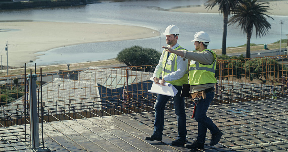 Builders reading a blueprint and talking at a construction site. Two men working as a team on an architecture project. Engineers and colleagues discussing plans