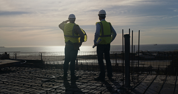 Two building contractors inspecting a construction site. Rear view of male colleagues standing on a building during an architecture project and looking at the view