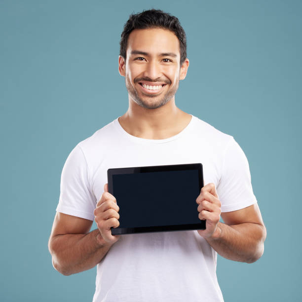 istock Handsome young mixed race man holding his digital tablet while standing in studio isolated against a blue background. Hispanic male showing you a website or product on his wireless device screen 1399365616