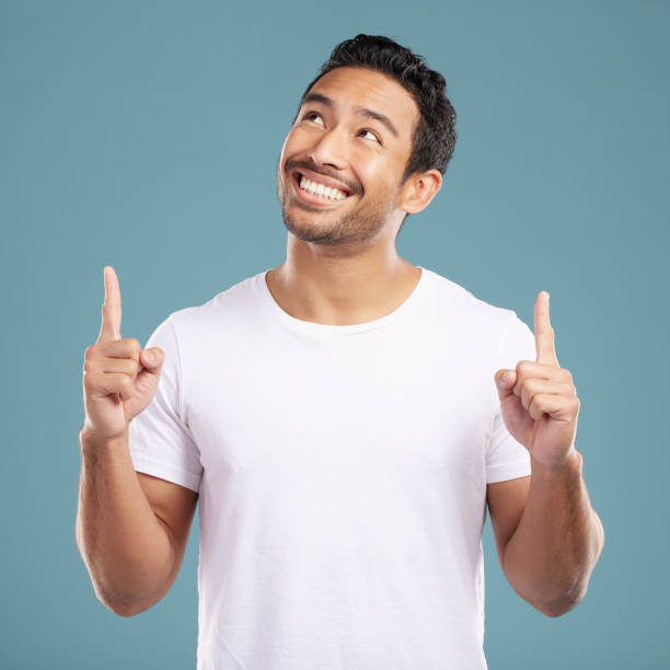 handsome young mixed race man pointing towards copyspace while standing in studio isolated against a blue background. happy hispanic male advertising or endorsing your product, company or idea - directly below imagens e fotografias de stock