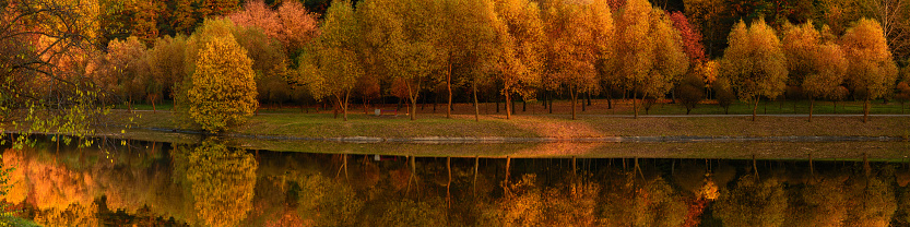 vibrant colors of october in beautiful autumn landscape. wide picturesque panoramic view of deserted orange evening city park along the coast on the with lush trees reflected in the water