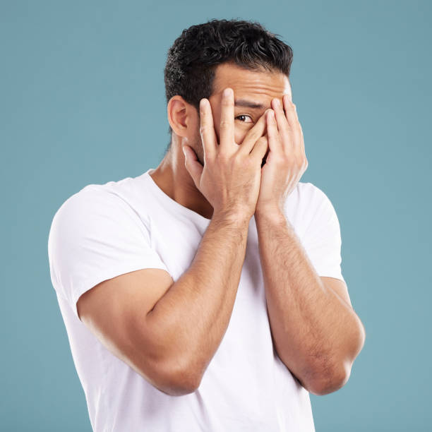 istock Handsome young mixed race man peaking through his fingers and covering his face with his hands while standing in studio isolated against a blue background. Hispanic male looking shy and hiding away 1399366108