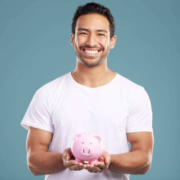 istock Handsome young mixed race man holding his pink piggybank while standing in studio isolated against a blue background. Hispanic male showing savings, finance, investment, wealth management and banking 1399366119