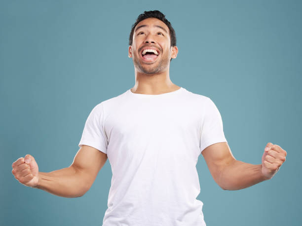 apuesto joven mestizo que celebra la victoria o el éxito mientras está de pie en el estudio aislado sobre un fondo azul. hombre hispano animando y bombeando sus puños ante el éxito o el logro - cheering men t shirt celebration fotografías e imágenes de stock