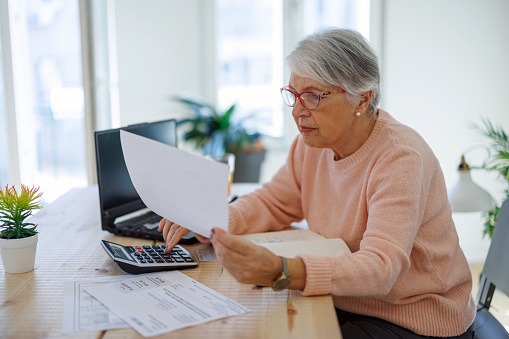 Senior caucasian woman checking pension documents, loan, e-banking, doing paperwork with laptop, mortgage, paying domestic bills at home
