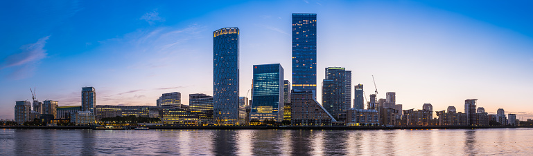The glittering lights of Canary Wharf Docklands financial district skyscrapers and modern apartment blocks reflecting in the tranquil waters of the River Thames before sunrise in the heart of London, the UK’s vibrant capital city.