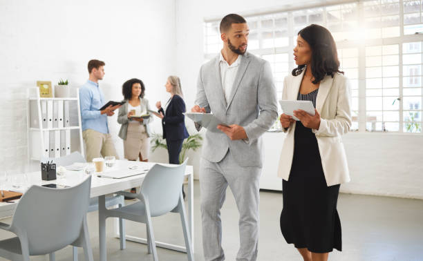istock Mixed race businessman and african american businesswoman talking while using a digital tablet in an office with coworkers. Businesspeople working together 1399369637