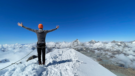 Magnificent view of a person on a snow-covered mountain summit of Breithorn in Switzerland. Celebrating a success. Looking down from the top with arms spread. Taking in the view