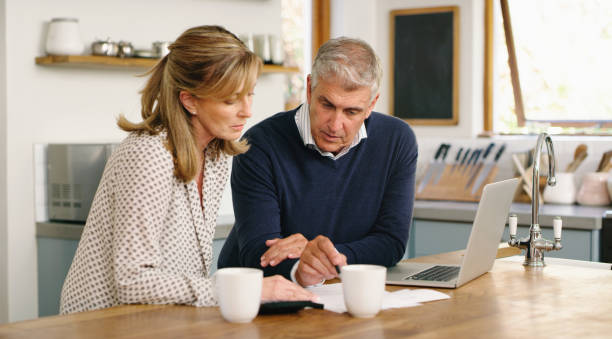 una pareja de personas mayores que planifica sus finanzas y paga facturas mientras usa una computadora portátil en casa. un hombre y una mujer maduros que pasan por el papeleo y trabajan en línea con una computadora - looking at papers fotografías e imágenes de stock