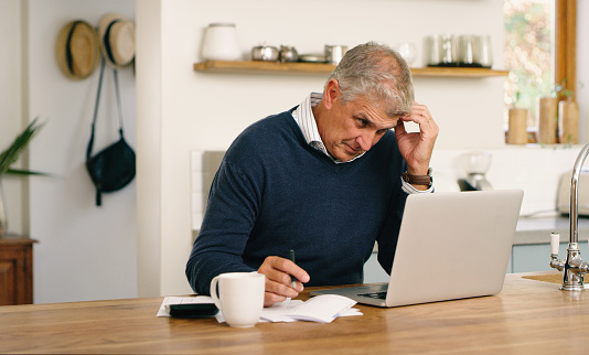 A senior man planning his finance, paying bills and looking unhappy while using laptop at home. A mature man going through paperwork and working online with a computer and feeling confused, stressed and unsure