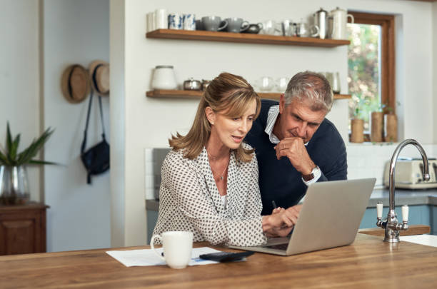 una pareja de personas mayores que planifica sus finanzas y paga facturas mientras usa una computadora portátil en casa. un hombre y una mujer maduros que pasan por el papeleo y trabajan en línea con una computadora - 50 59 años fotografías e imágenes de stock