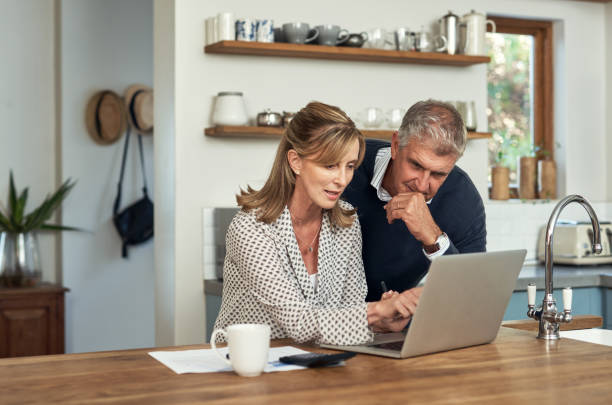 A senior couple planning their finance and paying bills while using a laptop at home. A mature man and woman going through paperwork and working online with a computer stock photo