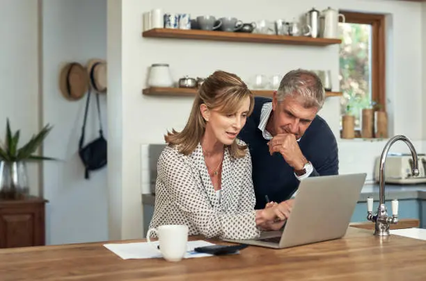 A senior couple planning their finance and paying bills while using a laptop at home. A mature man and woman going through paperwork and working online with a computer