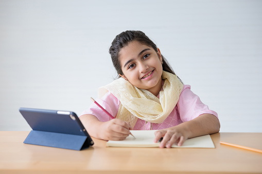 portrait cute Indian girl writing on notebook and online learning class from tablet, education concept