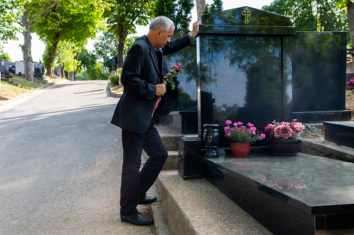 Mature man in black clothes on cemetery, holding a flower and mourning for family loss. Concept for death, mourning, funeral and spirituality.