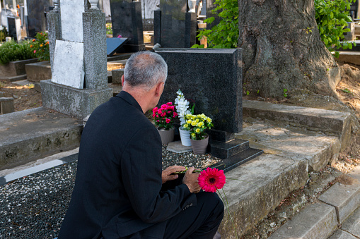 Mature man in black clothes on cemetery, holding a flower and mourning for family loss. Concept for death, mourning, funeral and spirituality.