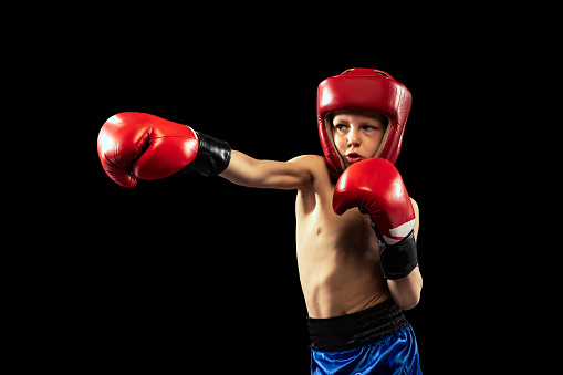 Right hook. Dynamic portrait of sportive boy, kid in boxer gloves and shorts practicing isolated on dark background. Concept of sport, movement, studying, achievements lifestyle. Copy space for ad