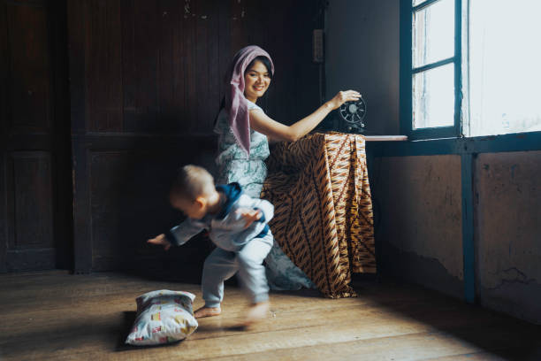 malay women stitching cloths by machine at home while her son playing beside her