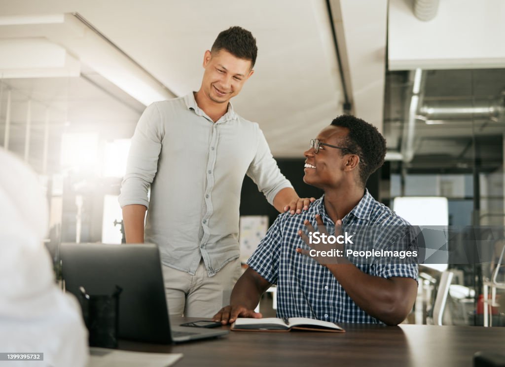 Two diverse businessmen talking at a desk discussing new project on laptop. Mentor complimenting colleague on a job well done and being a fast learner in modern office Trainee Stock Photo